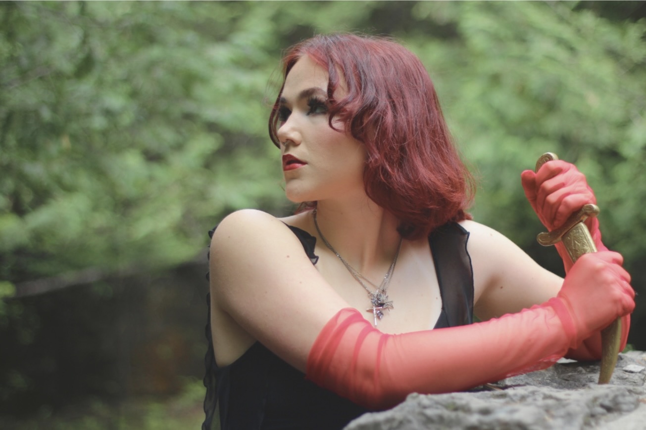 A person with red hair and red gloves stares into the distance in a forest. 