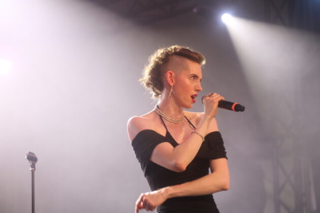 A woman in a black dress with red hair sings on a stage with white backlighting.
