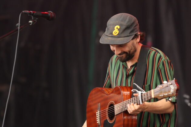 A man with a green stripped shirt and ball cap holds a guitar and looks at the ground.