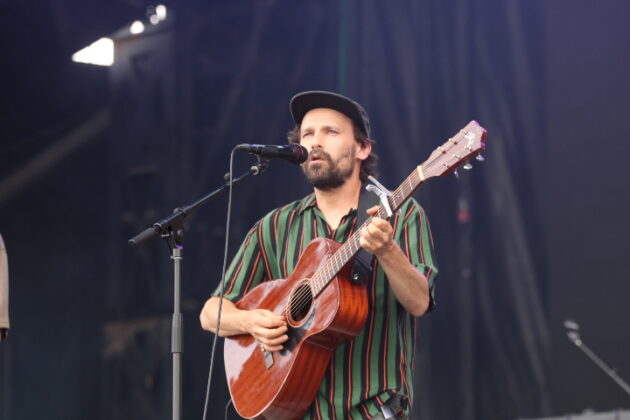A man with a baseball cap and a stripped green shirt plays guitar and sings on a stage.