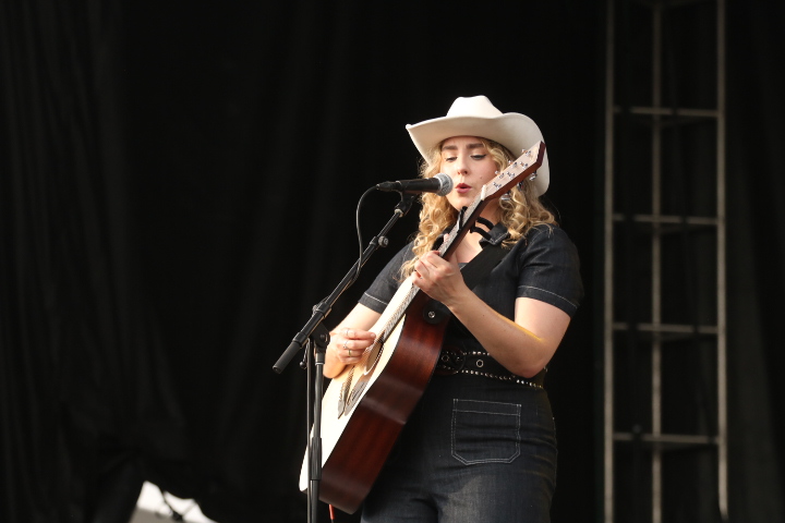 A woman with a white cowboy hat and a black outfit plays guitar and sings. 
