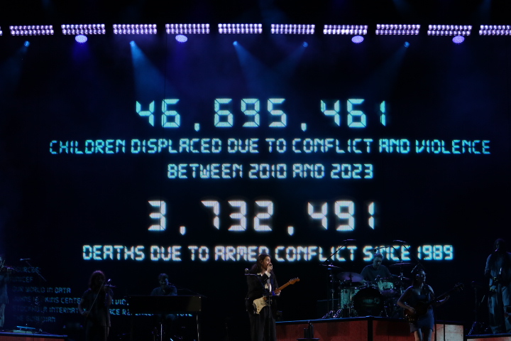 A man performs on a stage with a band as numbers of displaced children flash in the background.