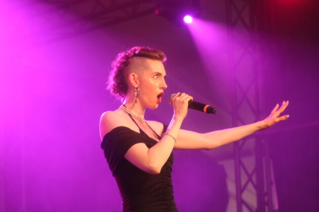 A woman with red hair in a black dress under a purple spotlight passionately sings.