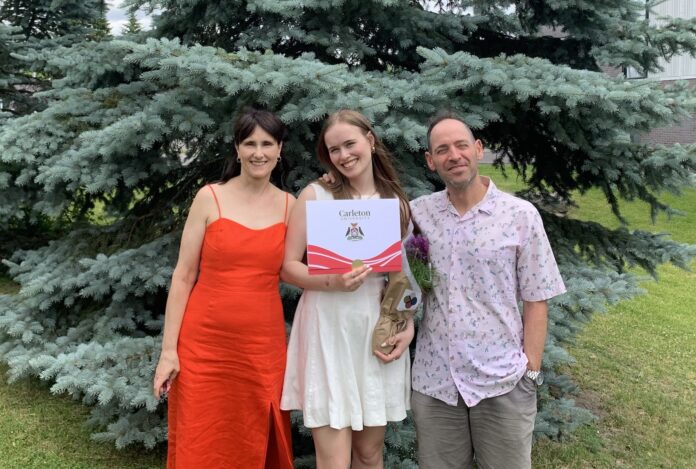 Three people stand in front of a large pine tree, the person in the middle holds up a graduation certificate.