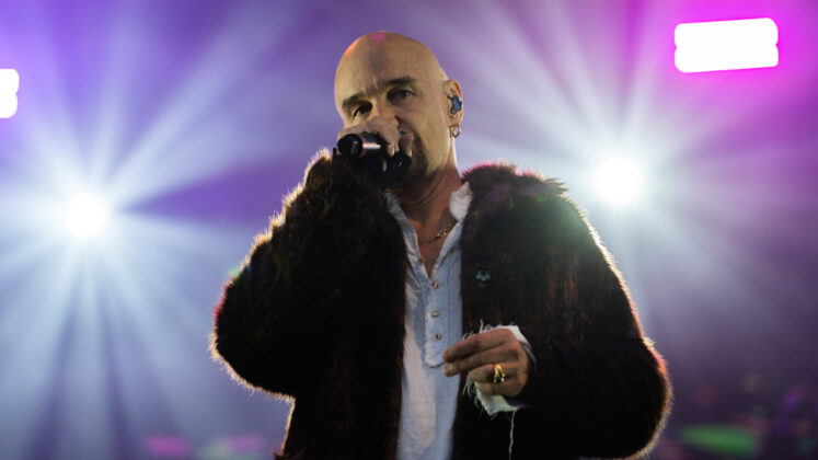 A singer in a brown fur jacket sings on stage in front of white spotlights.