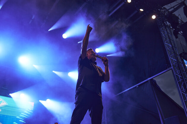 A performer sings with his arm pointed towards the sky on stage under blue lighting.