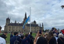 Ottawa raises Deaf flag at Parliament A blue and yellow flag is raised on a flagpole on Parliament Hill in front of a crowd with one person point their phone towards the flag.