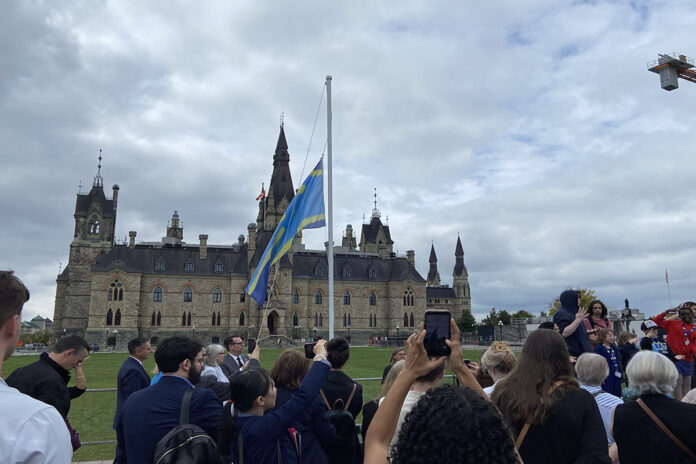 A blue and yellow flag is raised on a flagpole on Parliament Hill in front of a crowd with one person point their phone towards the flag.