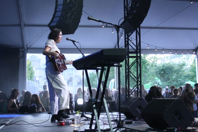 A singer in a white outfit with an acoustic guitar plays on stage in daylight.