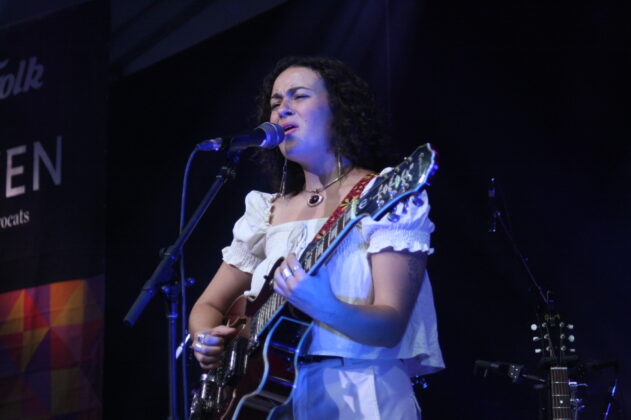 A singer in a white outfit with an acoustic guitar plays on stage in daylight.