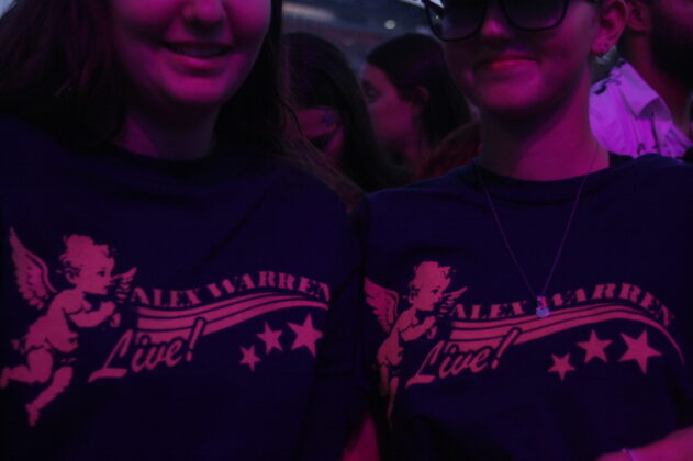 Two people stand wearing the same "Alex Warren" t-shirts at the barricade of a concert.