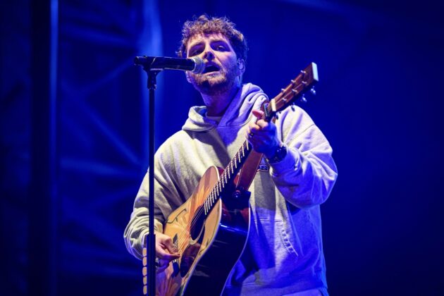 A singer performs on stage in a gray hoodie with an acoustic guitar