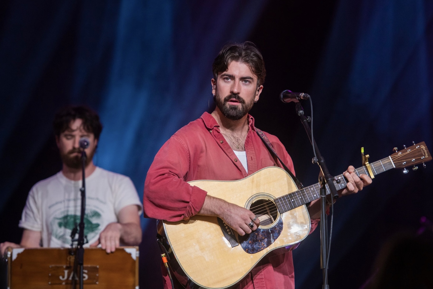 A band performs on stage, the lead singer has a red shirt and acoustic guitar.