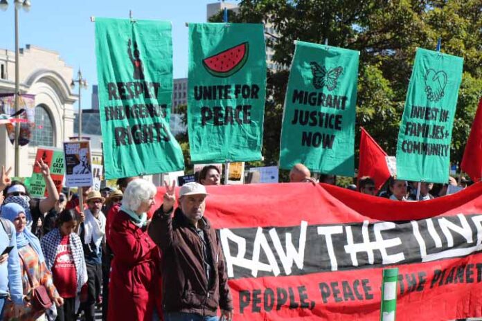 a crowd of people hold a wide red banner that says 'draw the line: people, peace, the planet'