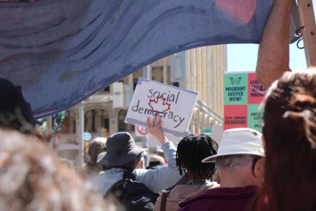 a crowd of people with somebody holding up a sign with a red maple leaf and the words, 'social democracy'