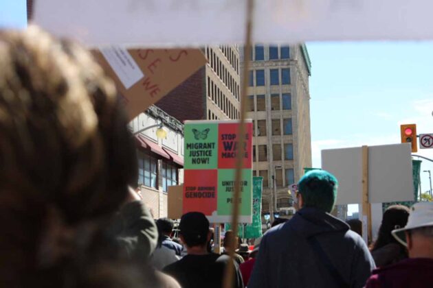 a crowd of people walk with one person holding up a sign saying 'migrant justice now,' 'stop the war machine,' 'stop arming genocide,' and 'united for justice and peace'
