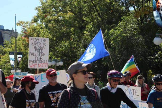 a crowd with a flags such as a pride flag and posters such as one that says 'from the river to the sea, free palestine for everyone! Why not?!'