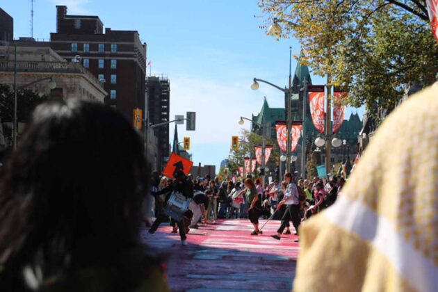 a crowd of people paint the cement on the street red with an unreadable message. People surround those painting, and someone drums.