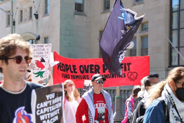 Group of people holdings walk past a building. One large banner is red and says 'people over profit, fund families + communities'
