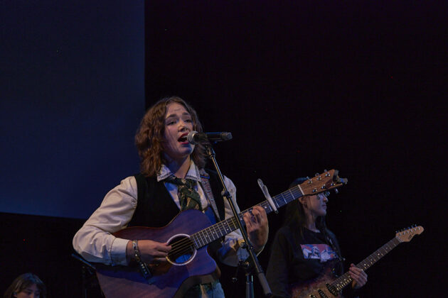 A person with an acoustic guitar sings to a crowd in a darkly-lit room.