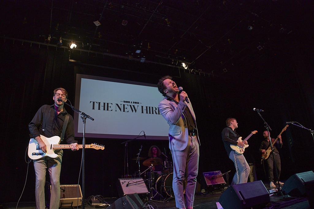 A band performs on a stage with the background reading "The New Hires."