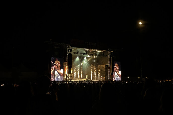 A singer in a white t-shirt sings on a stage lit up by yellow sparklers at night.