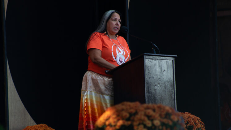 woman stands at podium wearing an orange shirt and an orange ribbon skirt