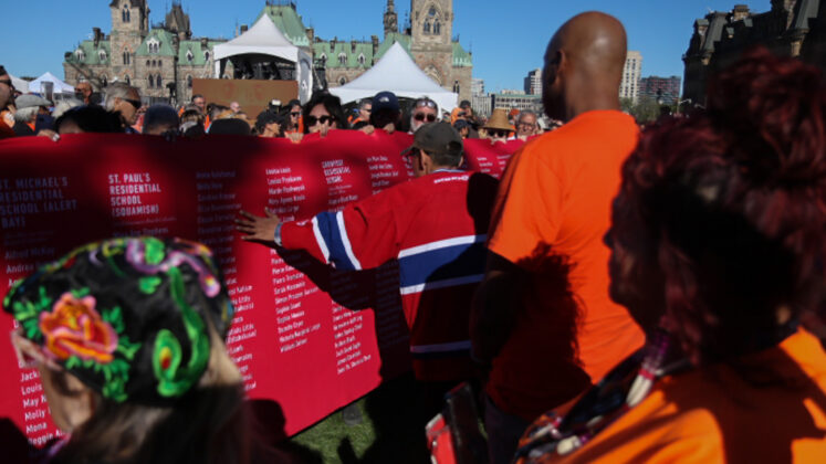 A group walks while holding up a long banner with a list of residential schools with children's names underneath.