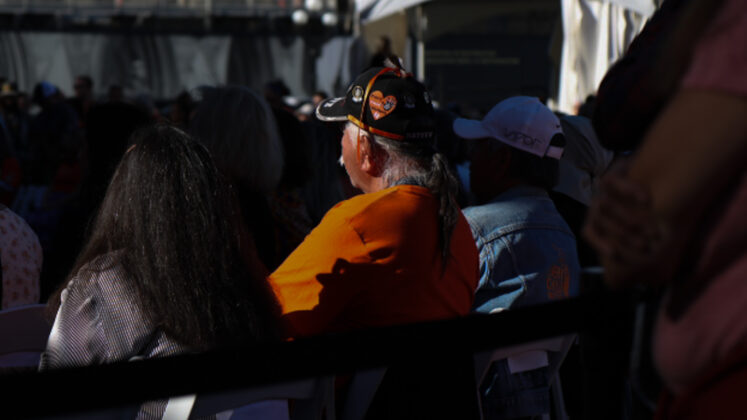 man sits in crowd wearing an orange shirt and a hat with an orange heart