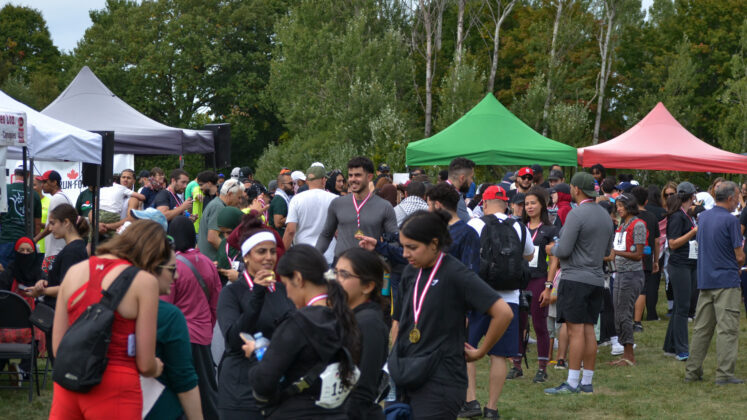 A crowd, with many wearing medals and atheltic wear talk to each other and smile with event tents nearby on the field.