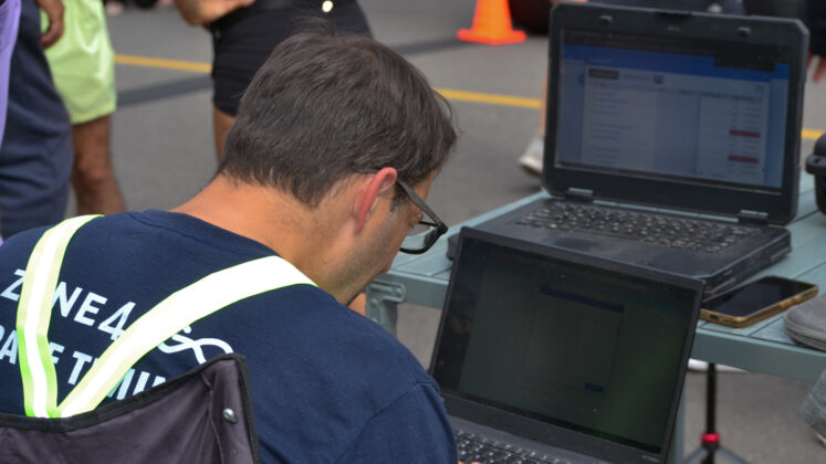 A man with glasses types on a laptop on his knees, while another laptop sits on a table in front of him.