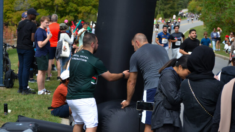 Two men hold up a black inflatable finish line as runners in the distance approach.