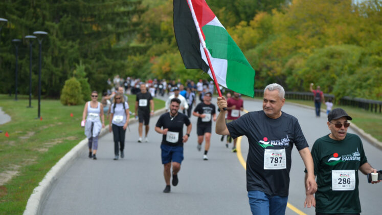 A man walks while waving a Palestinian flag. There are several runners behind him.