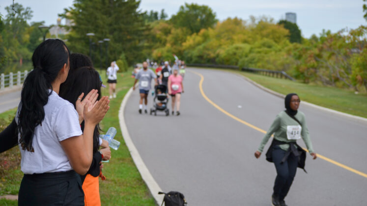 A woman surrounded by other people claps her hands from the sidelines as runners and walkers pass by.