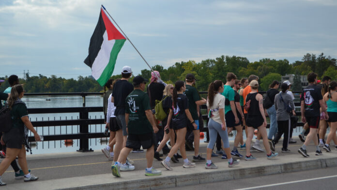 A group of people wearing athletic clothes walk on a bridge, with one person holding a Palestinian flag