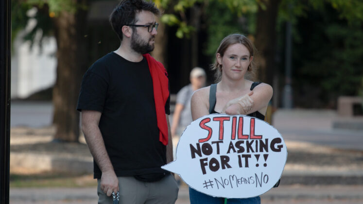 A person holds a sign shaped like a speech bubble with text saying "still not asking for it!!"