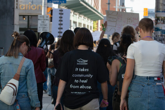 The backs of three people walking downtown. One wears a t-shirt saying "it takes a community to heal from abuse."