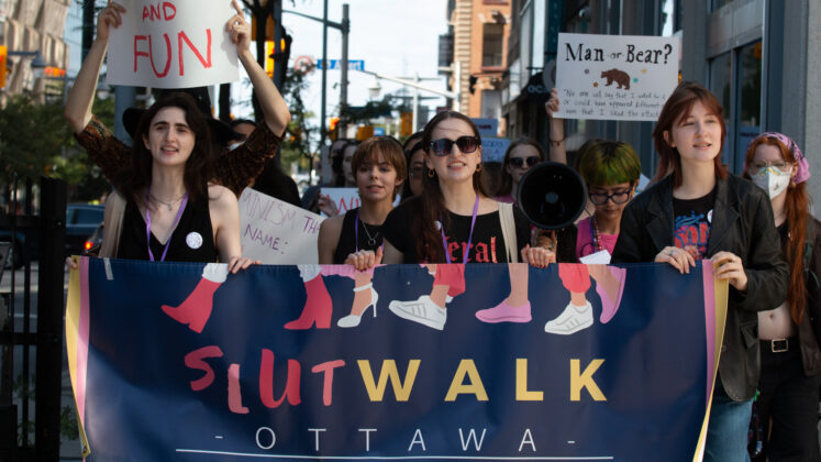 A crowd of people hold a banner with graphics of shoes walking and text that reads SlutWalk Ottawa