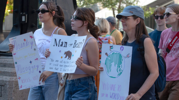 A group walks by a statue holding signs saying things like "man or bear" and "you'd have to stop the world just to skip the feeling"