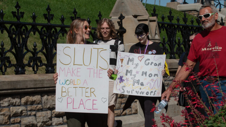 Four people walk by Parliament buildings, two of them holding signs. One sign reads "Sluts make the world a better place." The other sign reads "My gram, my mom, me, survivors"