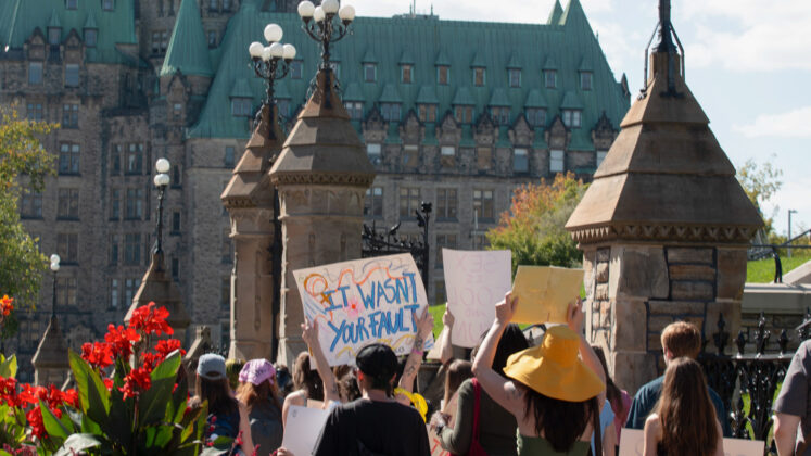 A crowd of people in front of a Parliament building with one person holding up a sign reading "It wasn't your fault"