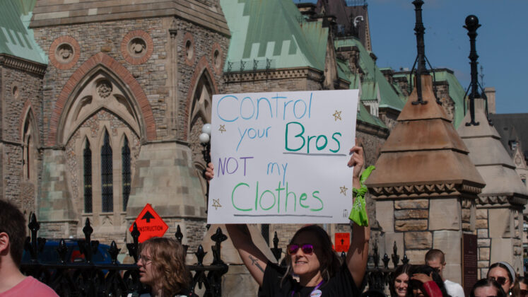 A group walks by Parliament buildings, with one person holding a sign saying "control your bros not my clothes"