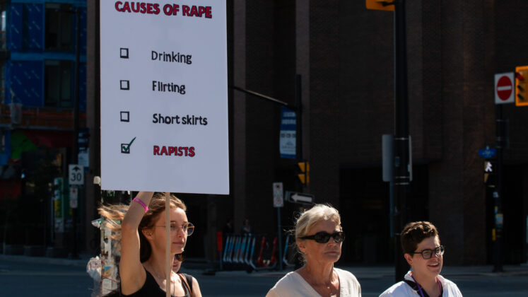 Three people walk on a street. One holds a sign with a checklist. The checklist title is "Causes of rape." The boxes are "drinking," "flirting," "short skirts," and "rapists." The latter is the only one checked off.