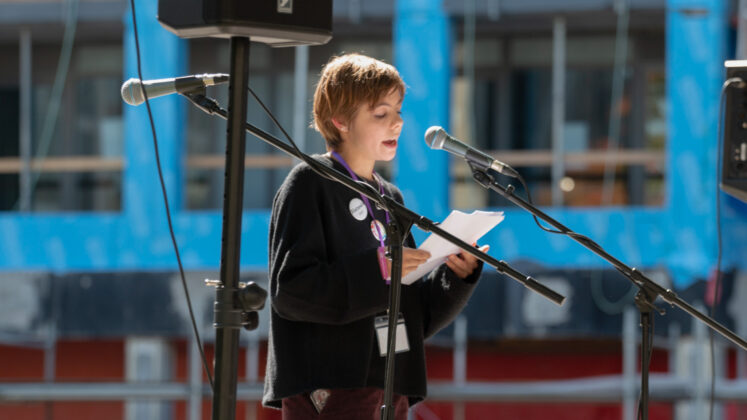 A person stands in front of a microphone and speaks while holding a paper in hand.