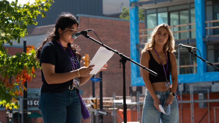 Two young women stand in front of microphones. One is speaking and looking at a paper in her hand.