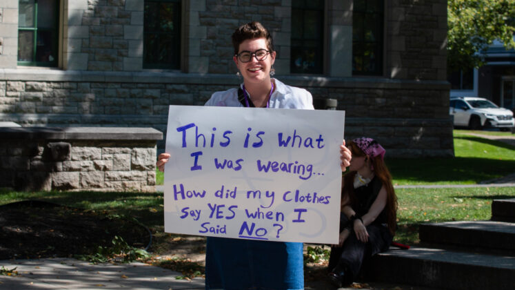 Person stands in front of city hall with a sign reading "This is what I was wearing... How did my clothes say yes when I said no?"