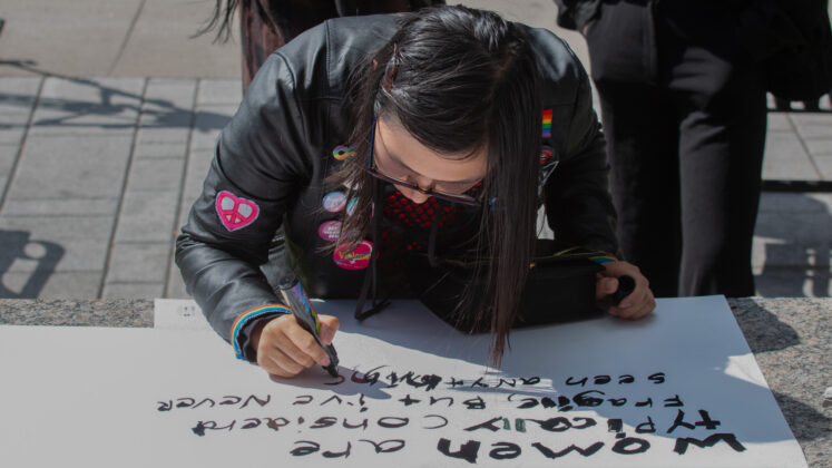 Person is bent over to write with a marker on a white poster saying "women are typically considered fragile but I've never seen anything..."