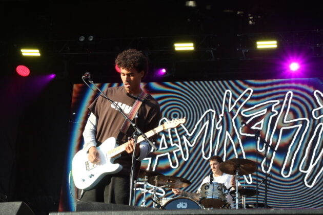 A singer performs on stage in daylight with an electric guitar.