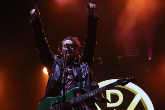 A guitarist sings and waves his hands on stage under orange lighting.