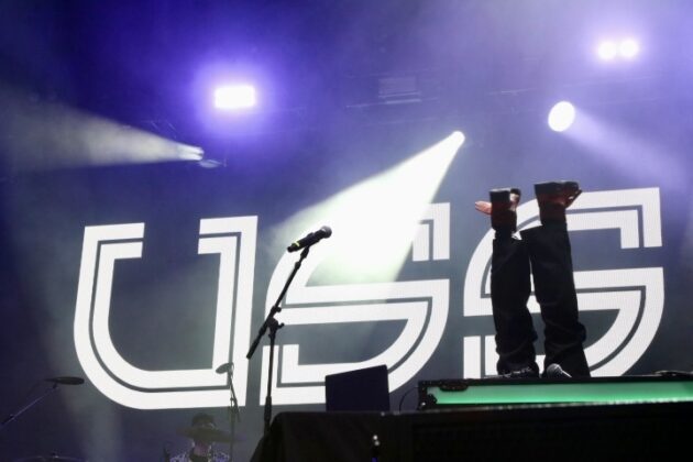 A band performs on stage under multi-coloured lighting.